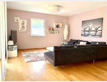 Wohnzimmer mit Holzboden, einer L-förmigen schwarzen Couch und einer rosa Wand. An der Wand hängen Familienfotos und ein großes Bild eines Flugzeugs, daneben stehen ein weißes Regal und eine Lampe. Ein kleiner Teppich liegt vor der Couch.