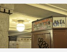 Ein U-Bahn-Zugang mit einer Wandverkleidung aus cremefarbenen Fliesen und einer hängenden Retro-Lampe an der Decke. Im Vordergrund ist ein brauner, mit Graffiti besprühter Zigarettenautomat mit einer oberen Beschilderung zu sehen.