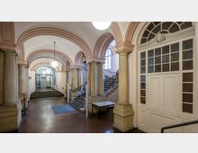 Flur eines historischen Gebäudes mit hohen, verzierten Rundbögen und massiven Säulen aus Stein. Eine breite Treppe mit schmiedeeisernem Geländer führt nach oben, und im Hintergrund sind Türen und eine Glastür sichtbar; die Decke ist mit Ornamenten geschmückt.
