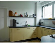 Helle Küchenzeile in einer L-Form mit cremefarbenen Schränken und schwarzer Arbeitsplatte. An der weißen, gefliesten Wand befinden sich eine Ablage mit Dekorationen und Gewürzen, darüber ein großes Fenster, das Tageslicht hereinlässt.