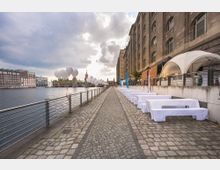 Gepflasterter Uferweg entlang eines Kanals mit Metallgeländer, gesäumt von einer historischen Backsteinfassade rechts und weiß gedeckten Biertischen. Links ist Wasser mit Gebäuden am gegenüberliegenden Ufer und der Oberbaumbrücke im Hintergrund zu sehen, dekoriert mit weißen Luftballons.