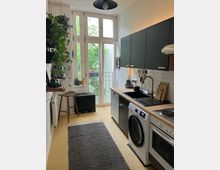 Schmale Küche mit einer Arbeitsfläche aus Holz, schwarzen Oberschränken und integrierten Geräten wie einer Waschmaschine und einem Herd. Helle Altbau-Doppelfenster, die zu einem kleinen Balkon mit grüner Aussicht führen, sowie großzügige Pflanzen und ein gemusterter Teppich prägen den Raum.