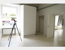 Heller Raum mit weiß gestrichenen Wänden und Fußboden, einem großen Fenster mit Heizkörper darunter und sichtbarer Verkabelung entlang der Wände. Im Vordergrund steht ein Stativ, im Hintergrund ist eine geöffnete Tür zu einem weiteren Raum sichtbar.