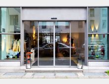 Schaufenster eines modernen Einzelhandelsgeschäfts mit großen Glasfronten, durch die ein einladender, beleuchteter Innenraum mit Displays und dekorativen Gegenständen zu sehen ist. Auf den Fenstern sind der Name „Le Magasin by Valise“ und Werbebotschaften aufgedruckt, und außen stehen dekorative Laternen.