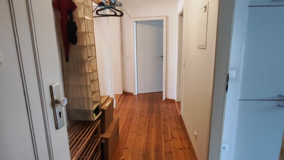 Narrow apartment hallway with white walls and a varnished wooden plank floor, leading to a closed door at the end. On the left are open coat hooks and a shoe rack, with doorways opening off the corridor on the right.