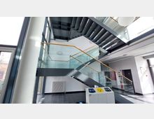 Innenbereich eines modernen Gebäudes mit einer schwarz-metallenen Treppe und Glasgeländern mit goldenen Handläufen. Der Raum hat helle Wände, große Fenster und auf dem Boden befinden sich dunkle Fliesen; im Vordergrund steht eine Recycling-Station.