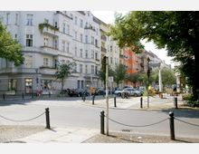 Straßenszene in einem Wohnviertel mit historischen Mehrfamilienhäusern, teils in weiß und orange gestrichen, und reich verzierten Fassaden. Im Vordergrund ein kleiner Platz mit Bäumen, parkenden Autos, Fahrrädern und einem Straßenschild; daneben sitzt eine Person auf der Terrasse eines Cafés.