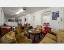 Gemütlicher Aufenthaltsraum mit verschiedenen gepolsterten Sesseln und Sofas um niedrige Holztische. Helle Wände mit Postern, ein Verkaufsautomat und ein angrenzender Raum mit schlichten Sitzgelegenheiten sind sichtbar.