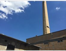 Fabrikgebäude mit einem hohen, gemauerten Schornstein, umgeben von flachen, beige-farbenen Gebäuden mit Wellblechdächern; darüber ein blauer Himmel mit vereinzelten Wolken.