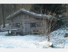 Holzhütte in einem verschneiten Waldgebiet mit moosbewachsenem Dach, kleinem Schornstein und zwei kleinen Fenstern. Vor der Hütte steht ein rustikaler Holztisch und ein kahler Baum; der Boden ist von einer dünnen Schneeschicht bedeckt.