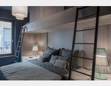 Schlafzimmer mit einem Hochbett aus Holz, das über zwei schwarze Leitern erreichbar ist. Darunter steht ein Doppelbett mit grauer Bettwäsche, flankiert von zwei Nachttischen mit grünen Tischlampen; ein Fenster befindet sich auf der linken Seite.