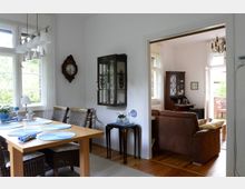 Essbereich in einem hellen Wohnraum mit einem Holztisch und geflochtenen Stühlen, gedeckt mit blauen Tellern. Im Hintergrund verbindet eine offene Tür den Essbereich mit einem Wohnzimmer, das mit einem braunen Ledersofa und Holzmöbeln eingerichtet ist; Parkettboden und weiße Wände prägen den Raum.