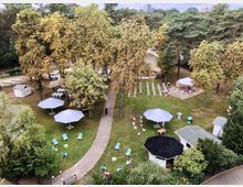 Luftaufnahme eines parkähnlichen Geländes mit großen Bäumen, Liegestühlen, weißen Sonnenschirmen und Sitzreihen, die auf eine kleine Bühne ausgerichtet sind. Im Hintergrund sind Gebäude und dichte grüne Vegetation zu sehen.