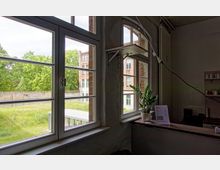 Innenraum mit Blick durch große Sprossenfenster auf eine grüne Rasenfläche, eine Backsteinmauer und ein mehrstöckiges Backsteingebäude mit weißen Fenstern; im Vordergrund ein Tisch mit einer Zimmerpflanze und einem laminierten Menü.