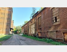 Industrielle Backsteinfassaden eines verlassenen Gebäudekomplexes, mit großen Fenstern und sichtbarer Abnutzung, neben einer schmalen gepflasterten Straße mit eingelassenen Gleisen. Im Hintergrund sind Bäume und ein klares Himmelblau sichtbar.