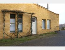 Gelbes Backsteingebäude mit einer weißen Eingangstür in Rundbogenform und mehreren rechteckigen Fenstern. An der Fassade ranken kahle Pflanzen über ein Metallgerüst, der Boden davor ist mit Gras und Asphalt bedeckt.