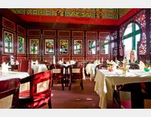Ein traditionell eingerichtetes Restaurant im asiatischen Stil mit dunklen Holzmöbeln, kunstvoll gestalteten Wänden mit floralen Mustern und reichlich verzierten Elementen. Weiße Tischdecken, gefaltete Stoffservietten und dekorative Vasen ergänzen das Interieur; große Fenster lassen Tageslicht einfallen.