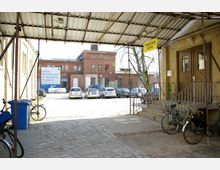 Hof eines ehemaligen Industriegebäudes aus rotem Backstein, mit abgestellten Autos und Fahrrädern. Rechts führt eine Treppe unter einem Überdachungskonstrukt zu einem Eingang mit Holzflügeltüren, darüber ein Schild mit der Aufschrift „Theater Café“.