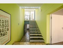 Treppenhaus mit hellgrünen Wänden, moderner Metalltreppe und Glasdecke, durch die Tageslicht einfällt. Im Vordergrund ein Steinboden, seitlich ein Plakat, oben ein Fenster mit Blick auf Bäume.