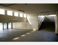 Unterführungsbereich mit Treppenaufgang und einem Ausgang mit Gittertüren. Wände und Decke sind mit weißen Fliesen verkleidet, und ein Hinweisschild mit Straßennamen ist sichtbar.