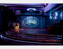 Ein dunkler Innenraum einer Event- oder Tanzfläche mit abgerundeter, holzverkleideter Bühne und LED-Lichtstreifen. Im Hintergrund befindet sich eine dekorierte Wand mit projiziertem Wolkenmotiv und Zahnräder als Wandschmuck.