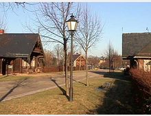 Ländliche Straße mit Holzhäusern auf beiden Seiten, Bäumen ohne Laub und einer schwarzen Straßenlaterne im Vordergrund. Die Umgebung ist von Rasenflächen und Hecken gesäumt.