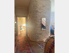 Wohnraum mit gewölbter Backsteinwand und Holzboden, ergänzt durch bunte Teppiche im orientalischen Stil. Im Hintergrund ist eine geöffnete Tür zu sehen, die zu einem hellen Raum mit Pflanzen und Holztreppe führt.