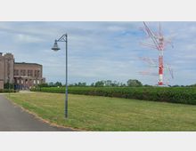 Backsteinbau mit großen Fenstern auf der linken Seite, daneben ein gepflasterter Weg und eine grüne Rasenfläche mit Hecken. Im Hintergrund steht ein großer, rot-weißer Sendemast vor einem Himmel mit leichter Bewölkung.