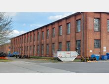 Backstein-Gebäude im Industriegelände mit hohen, regelmäßigen Fenstern. Im Vordergrund befinden sich Container, Bahnräder und verschiedene Metallteile auf einem betonierten Hof.
