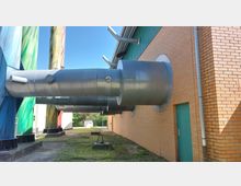 Außenansicht eines Industriegebäudes mit gelber Ziegelfassade, an der große metallische Lüftungsrohre herausragen. Im Hintergrund sind farbig bemalte Tanks und ein grüner Metallbereich des Gebäudes zu sehen.