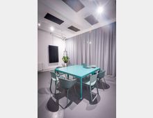 Moderner Besprechungsraum mit türkisfarbenem Tisch auf Rollen und mehreren passenden Stühlen auf grauem Boden. An der Decke hängen schallabsorbierende Paneele und Strahler, an der Wand befinden sich ein schwarzes Paneel sowie eine große graue Vorhangfront und eine hängende Pflanze.