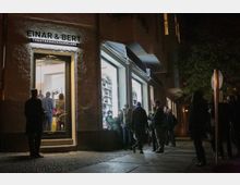 Außenansicht einer beleuchteten Theaterbuchhandlung bei Nacht mit der Aufschrift „Einar & Bert“. Mehrere Personen stehen vor dem Eingang und den großen Schaufenstern, in denen Bücher und Regale erkennbar sind.