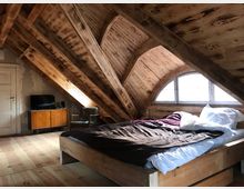 Dachgeschosszimmer mit Holzverkleidung und sichtbaren Balken, ausgestattet mit einem Bett, einem Sessel, einem kleinen Holzschrank und einem Fernseher. Eine halbrunde Fensterfront lässt Tageslicht herein.