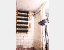 Ein kleiner, heruntergekommener Raum mit weiß gekachelten Wänden, teilweise beschädigt und stark verschmutzt. An der Wand befinden sich mehrere übereinander angeordnete Heizrippen mit rostroten Rohren, daneben ein Stativ mit einer Fotolampe.