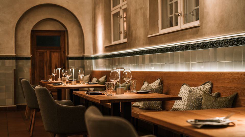 Gemütlicher Restaurant-Innenraum mit Holz­tischen, gepolsterter Sitzbank mit Kissen und Stühlen, jeweils mit Gläsern und Besteck gedeckt. Helle Putzwände mit Fliesenbordüre, hohe Fenster und ein Rundbogen mit Holztür prägen den Raum.
