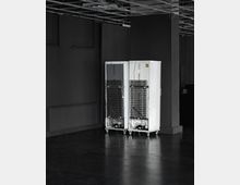 Zwei weiße Kühlschränke mit sichtbaren Lüftungsgittern auf der Rückseite stehen nebeneinander in einem dunklen Innenraum mit glattem Boden, Säulen und einer abgehängten Decke mit offenen Kabeln und Beleuchtungsschienen.