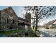 Historische Kirche aus Feld- und Ziegelsteinen mit einem steilen Satteldach und einem hohen Turm, umgeben von Bäumen und Grünflächen. Rechts der Kirche führt eine gepflasterte Straße vorbei, gesäumt von Fachwerkhäusern.