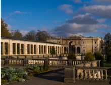 Herrenhaus mit klassizistischer Architektur, bestehend aus einer langen Glasgalerie mit Bogenfenstern und einer Fassade mit Säulen und Balustraden; umgeben von gepflegten Gartenanlagen und Bäumen unter einem bewölkten Himmel.