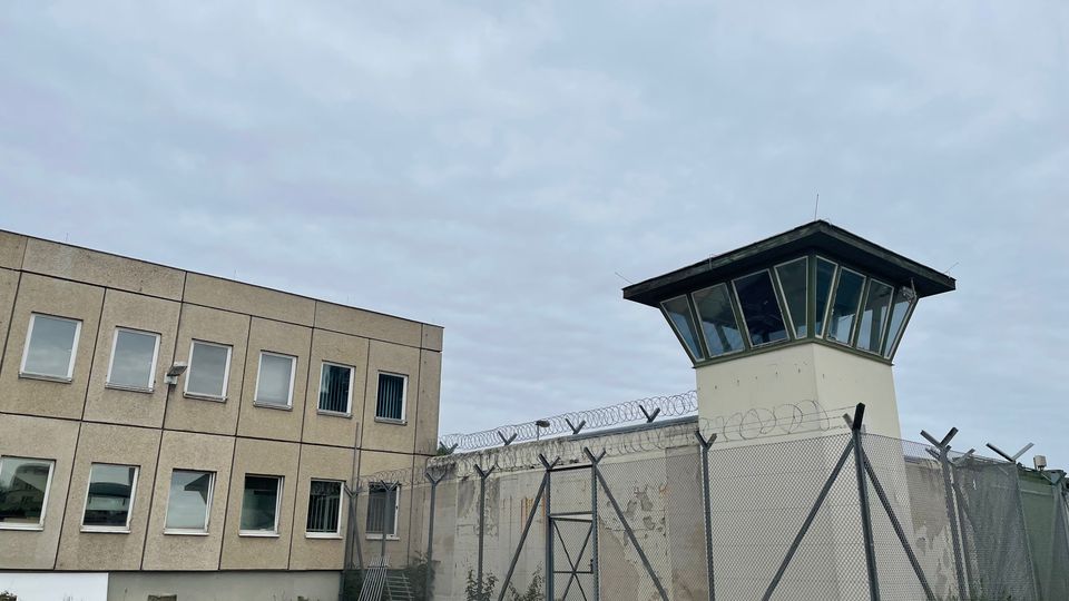 Außenansicht eines umzäunten Gefängnisareals mit hohem Maschendrahtzaun und Stacheldraht, daneben ein Wachtturm mit verglaster Aussichtsplattform. Links steht ein mehrstöckiges, rechteckiges Gebäude mit vielen Fenstern unter bewölktem Himmel.