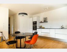 Moderne Wohnküche mit hellem Holzboden, weißen Einbauküchenschränken und integrierten Elektrogeräten. Im Vordergrund steht ein runder schwarzer Esstisch mit verschiedenen Stühlen, darunter ein roter und ein Drahtstuhl; an der Wand ein schwebendes Regal mit Dekoration.