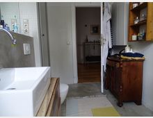 Badezimmer mit modernen und rustikalen Elementen: Ein weißes Waschbecken auf einem Holzunterschrank, eine Toilette und eine Dusche mit Glaswand; rechts ein hölzernes Sideboard und ein Wandregal mit Pflegeprodukten. Eine offene Tür führt in einen angrenzenden Raum mit Holzboden.