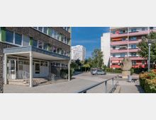 Mehrstöckige Wohngebäude in einer modernen Wohnsiedlung mit farbigen Fassaden und Balkonen; im Vordergrund ein gepflasterter Weg, eine Skulptur aus Stein und ein Parkplatz mit einem geparkten Auto, umgeben von Bäumen und Sträuchern.