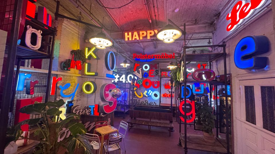 Ein Innenraum mit zahlreichen bunten, leuchtenden Buchstaben und Schriftzügen an den Wänden, Regalen und Decken. Der Raum hat eine industrielle Ästhetik mit unverputzten Wänden, Metallregalen und gedecktem Licht.