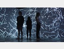 Drei Personen stehen in einem dunklen Raum vor einer interaktiven, beleuchteten Wand, die mit leuchtenden, abstrakten Linien und Punkten in Blau- und Weißtönen gestaltet ist. Der Boden reflektiert die grafischen Elemente der Wand.