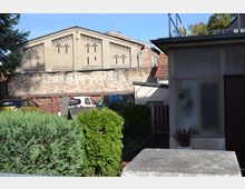 Hinterhof mit einer alten, teilweise verputzten Ziegelmauer und einem Gebäude mit auffälligen, dekorativen Lüftungsöffnungen. Im Vordergrund befinden sich Sträucher, parkende Autos und ein moderner Anbau mit Milchglasfenstern.