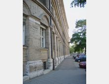 Straßenansicht eines historischen Gebäudes mit hellen Steinmauern, Rundbögen über den Fenstern und verzierter Fassade. Rechts des Gebäudes führt ein Bürgersteig mit parkenden Autos und vereinzelten Bäumen entlang.