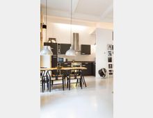 Open-plan modern kitchen and dining area with matte black cabinets, stainless steel range hood, and white tiled backsplash. A long wooden table with black chairs sits under two large silver pendant lights, with high ceilings and a white floor in a minimalist loft-style space.
