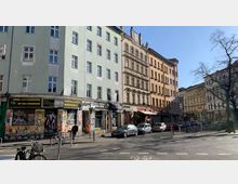 Wohnhäuserreihe in einer städtischen Straße mit mehrstöckigen Altbauten in unterschiedlichen Pastellfarben. Im Erdgeschoss befinden sich Geschäfte mit Graffiti und Plakaten an den Fassaden, davor parkende Autos und Fahrräder sowie eine belebte Straßenkreuzung.