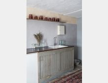 Rustikale Küche mit einer grauen Arbeitsfläche und einer Spüle, darunter eine Holzkonstruktion mit verwitterter Optik. Im Hintergrund eine graue Wand mit einem Wandregal, auf dem Gläser mit Eingemachtem stehen, sowie ein kleiner Hängeschrank. Ein Teppich mit geometrischem Muster bedeckt den Boden.