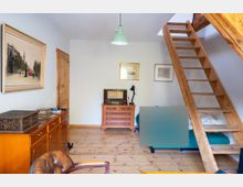 Wohnraum mit Holzfußboden, Kommode und gerahmten Bildern an hellen Wänden, beleuchtet von einer grünen Deckenlampe. Rechts führt eine offene Holztreppe zu einer Empore, darunter steht ein Bett oder Sofa.